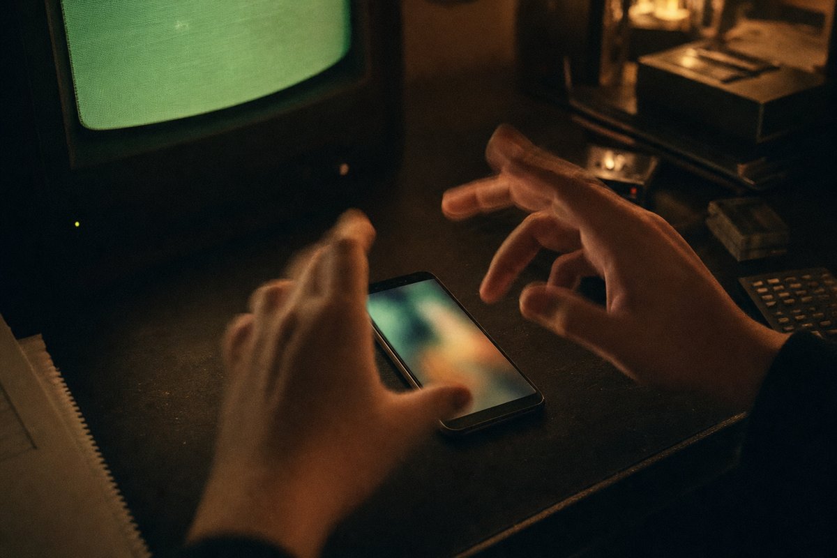 Due mani che compiono un gesto rapido su uno smartphone appoggiato su una scrivania scura, foto con bagliore verde-ambra